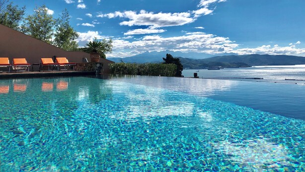 Una piscina infinita con agua azul cristalina, un área de descanso con sillones naranjas y una vista de montañas y una masa de agua al fondo.