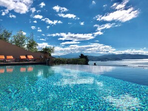 An infinity pool with clear blue water, a lounge area with orange chairs, and a view of mountains and a body of water in the background. - Mira-mare Apartments & Suites (La Spezia)