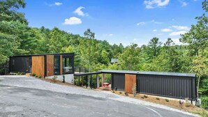 A modern cabin with a wooden deck, a covered outdoor seating area, and a view of a forested hillside. - Luxury Container | Retreat In Bloom • Pool • Sauna (Young Harris)