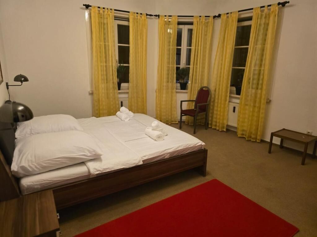 Una cama bien hecha con sábanas blancas, un tapete rojo y una silla en un rincón de una habitación con cortinas amarillas.