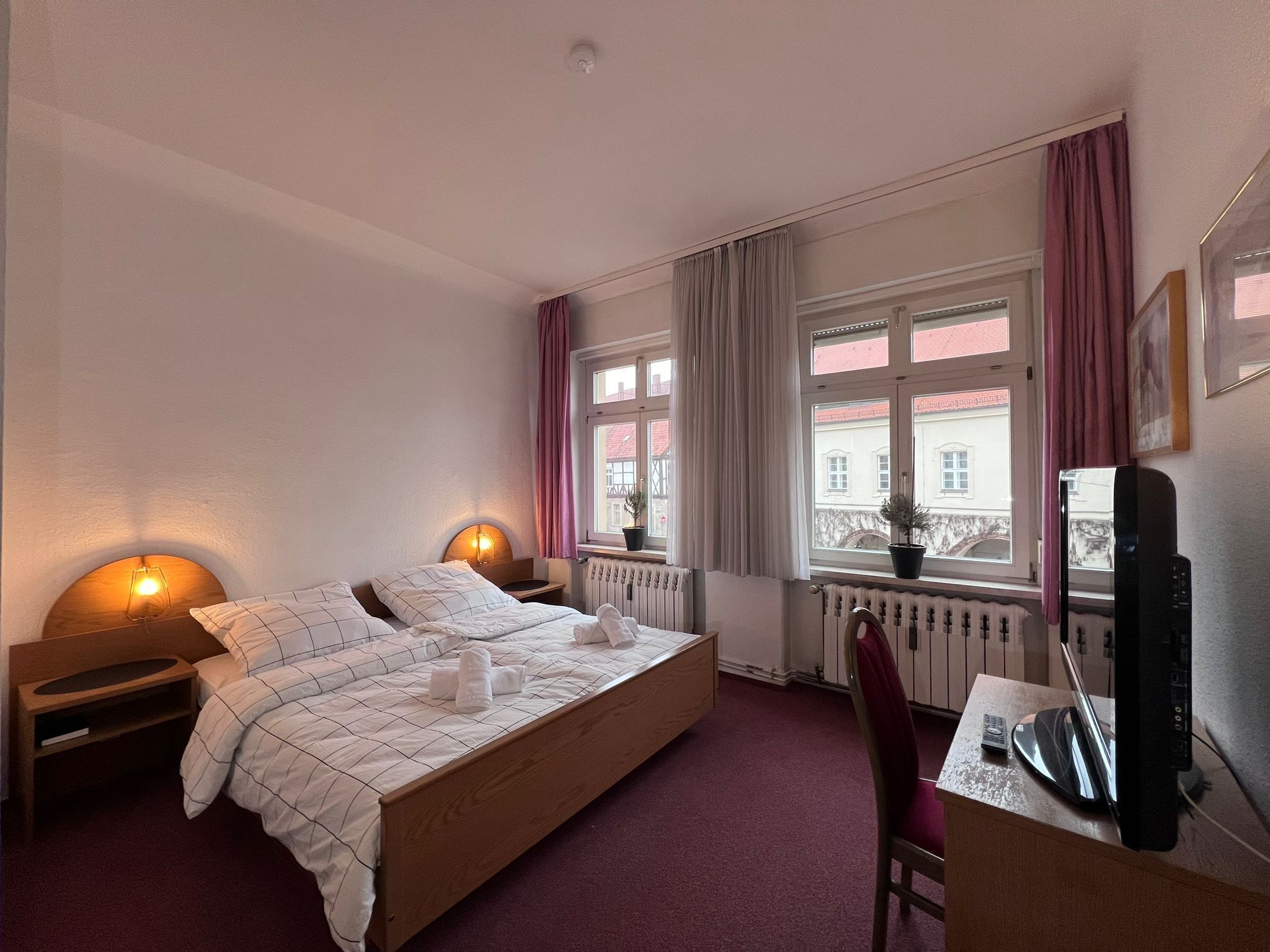 Ein Hotelzimmer mit Bett, Nachttischlampen, einem Schreibtisch mit Stuhl, einem Fenster mit Vorhängen und einer Heizung.