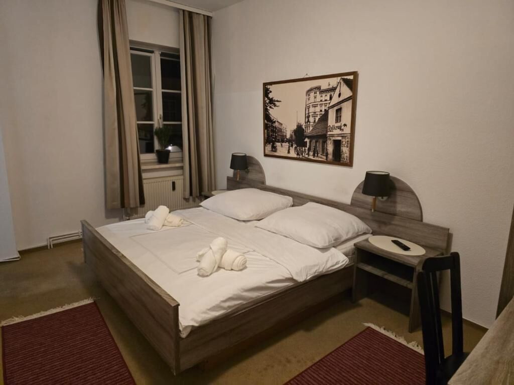Una cama bien hecha con sábanas y toallas blancas, una mesita de noche con lámpara, un cuadro en la pared y una ventana con cortinas.