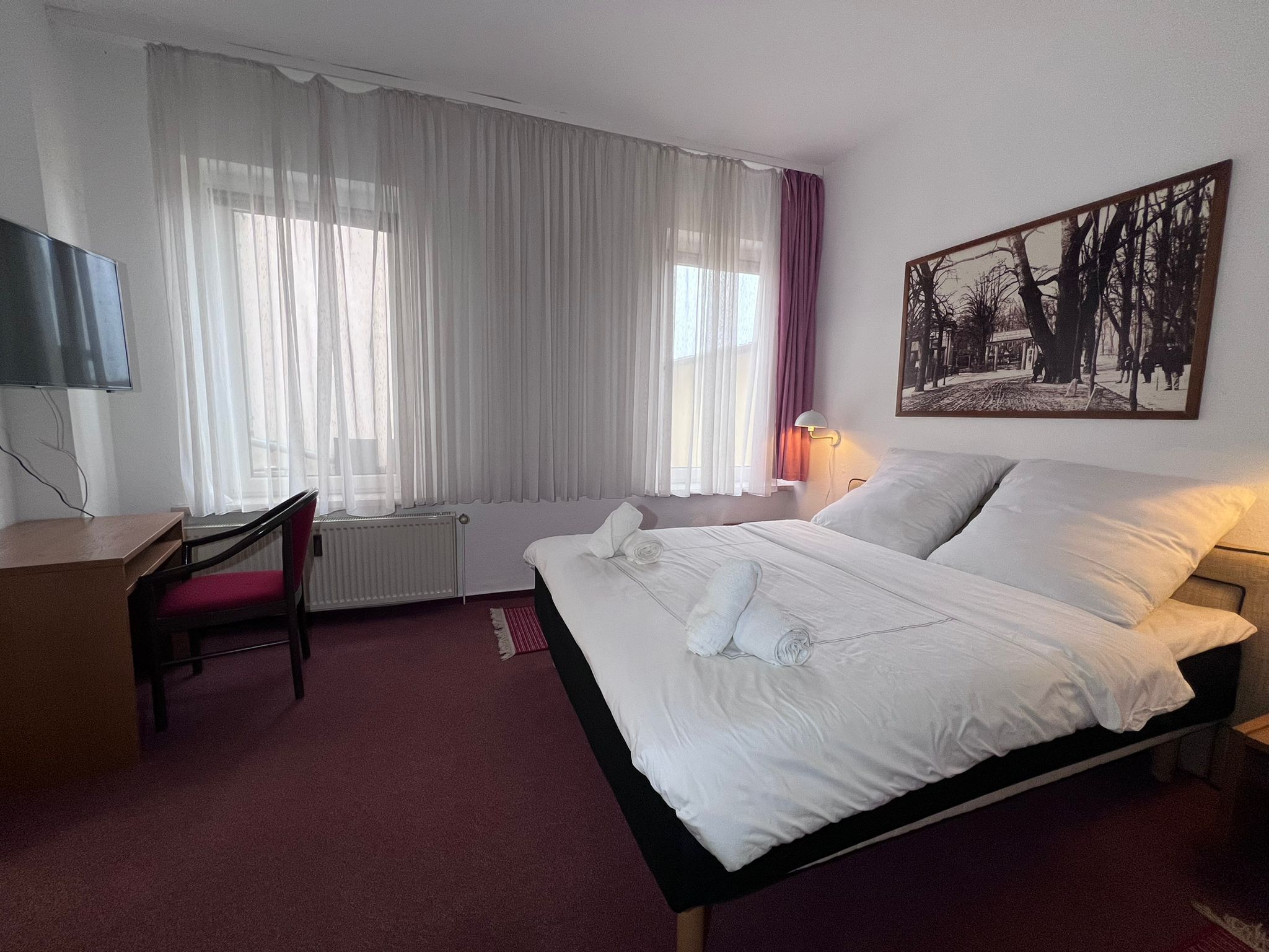 A hotel room with a bed, a desk, a chair, a radiator, a TV, and a painting on the wall.