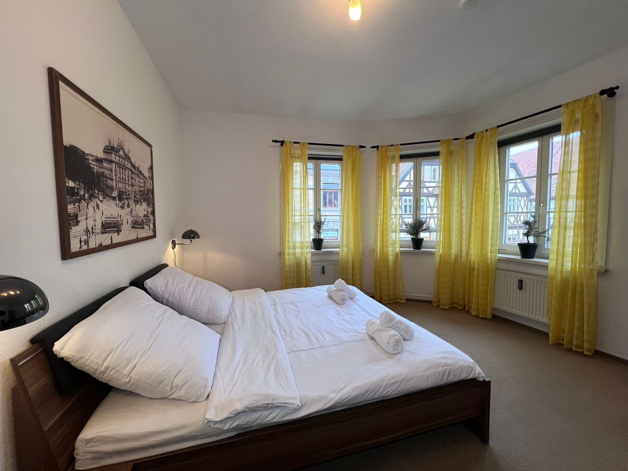 A hotel room with a large bed, a nightstand, a lamp, a framed picture on the wall, and a window with yellow curtains.