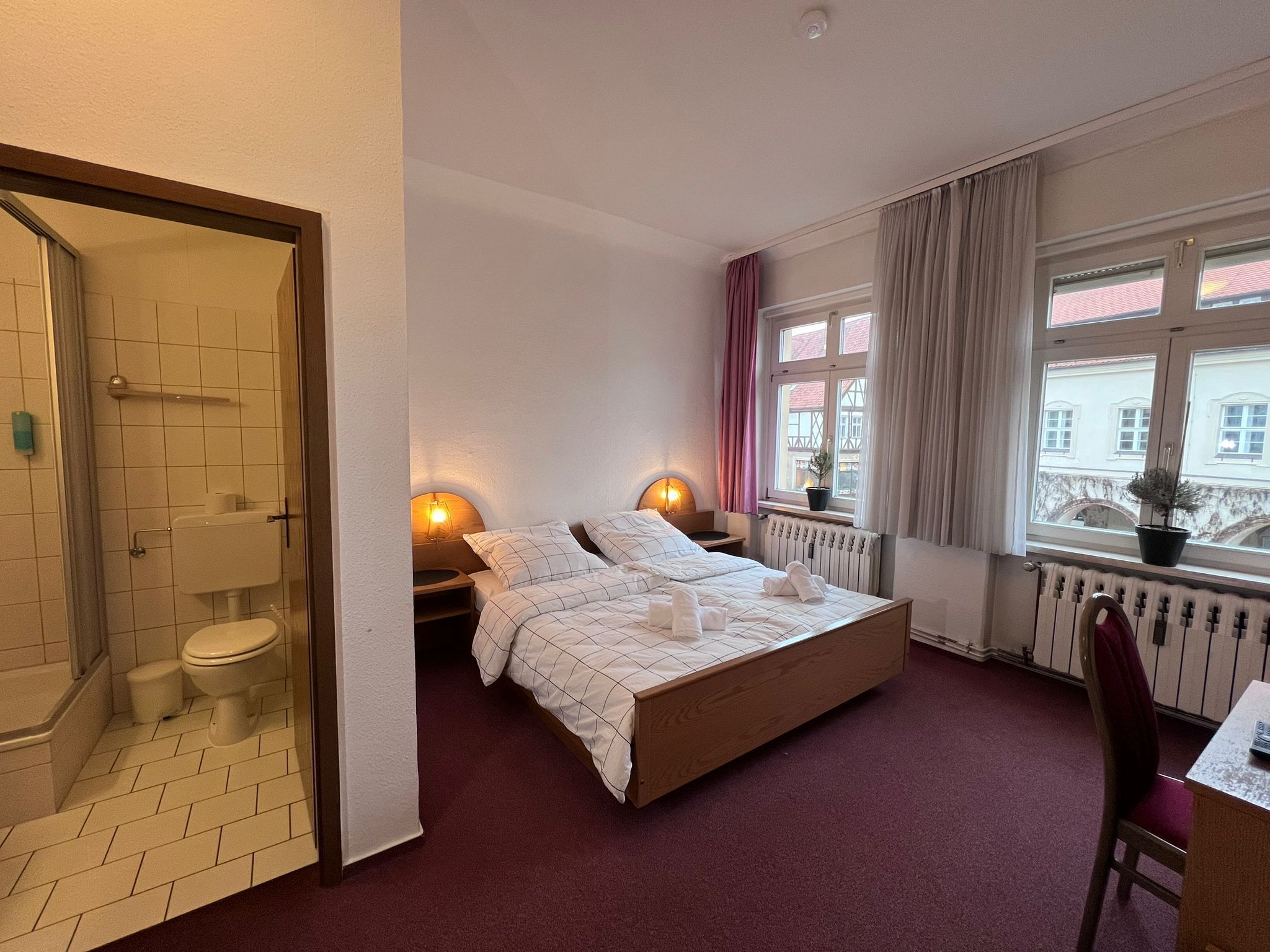 Ett hotellrum med en säng, ett skrivbord, en stol, ett fönster med gardiner, ett element och ett badrum med dusch och toalett.