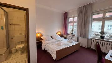 Ett hotellrum med en säng, ett skrivbord, en stol, ett fönster med gardiner, ett element och ett badrum med dusch och toalett.