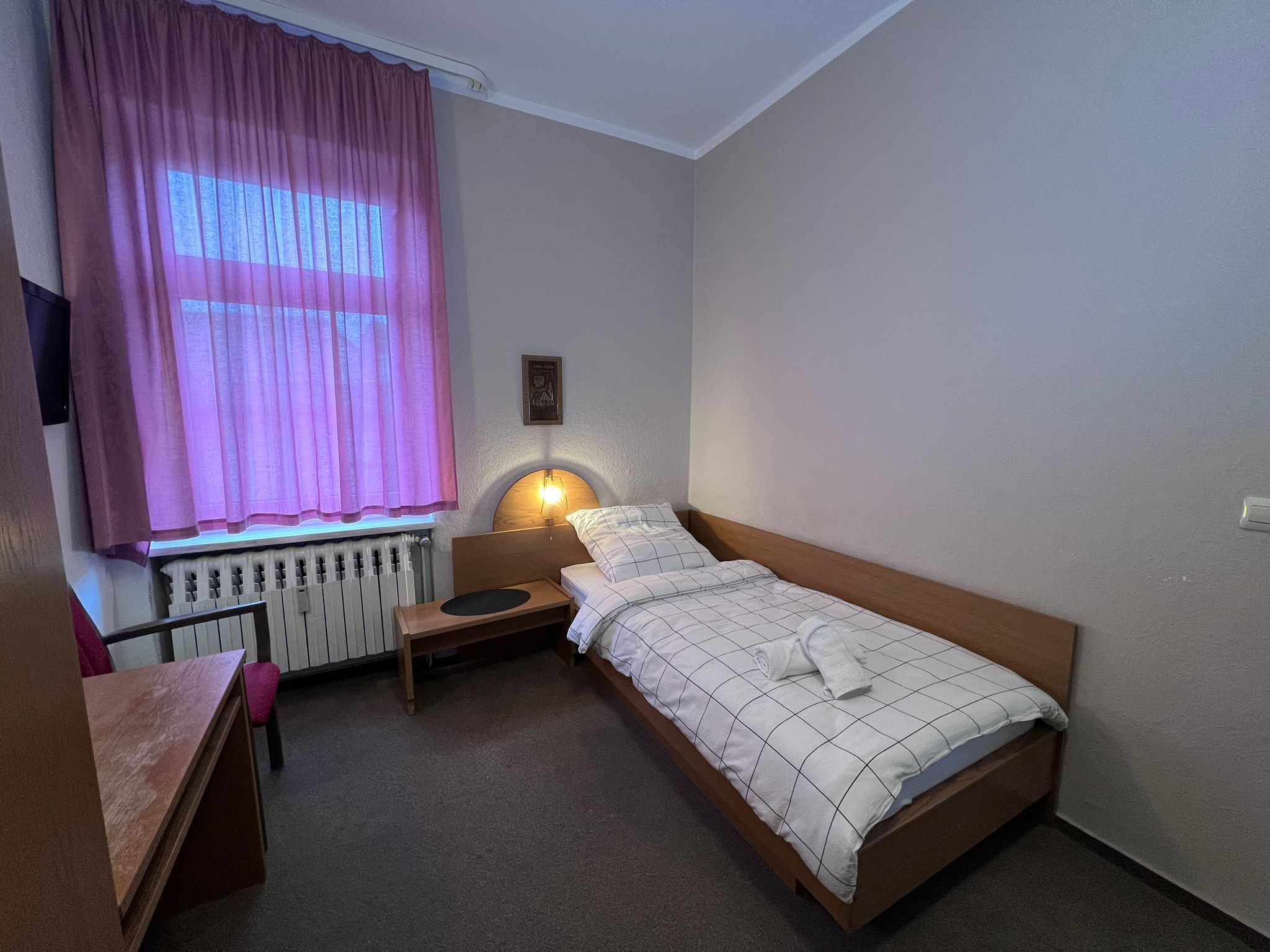 Un llit individual amb capçalera, una taula de nit, una làmpada, una finestra amb cortines i un radiador.