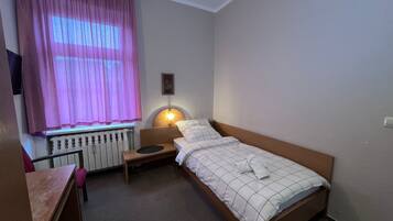 A single bed with a headboard, a bedside table, a lamp, a window with curtains, and a radiator.