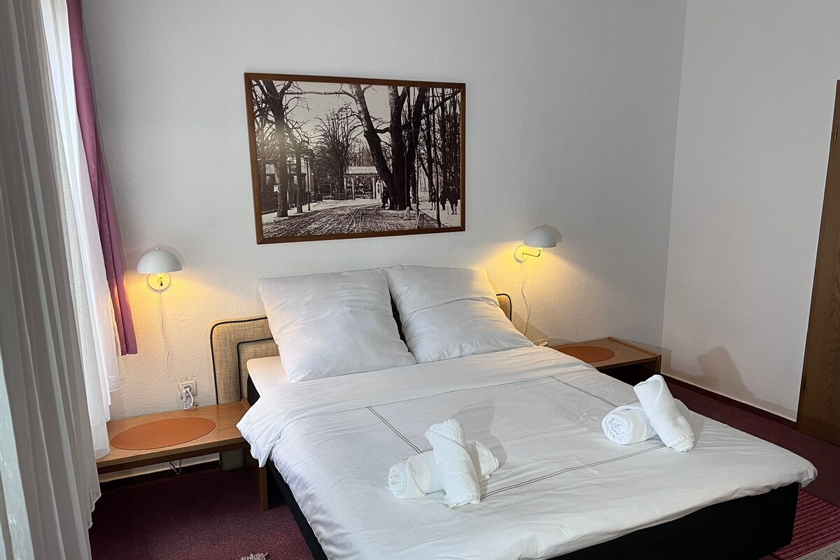Una cama bien tendida con sábanas y toallas blancas, dos lámparas de noche, un cuadro en la pared y una mesita de noche de madera.