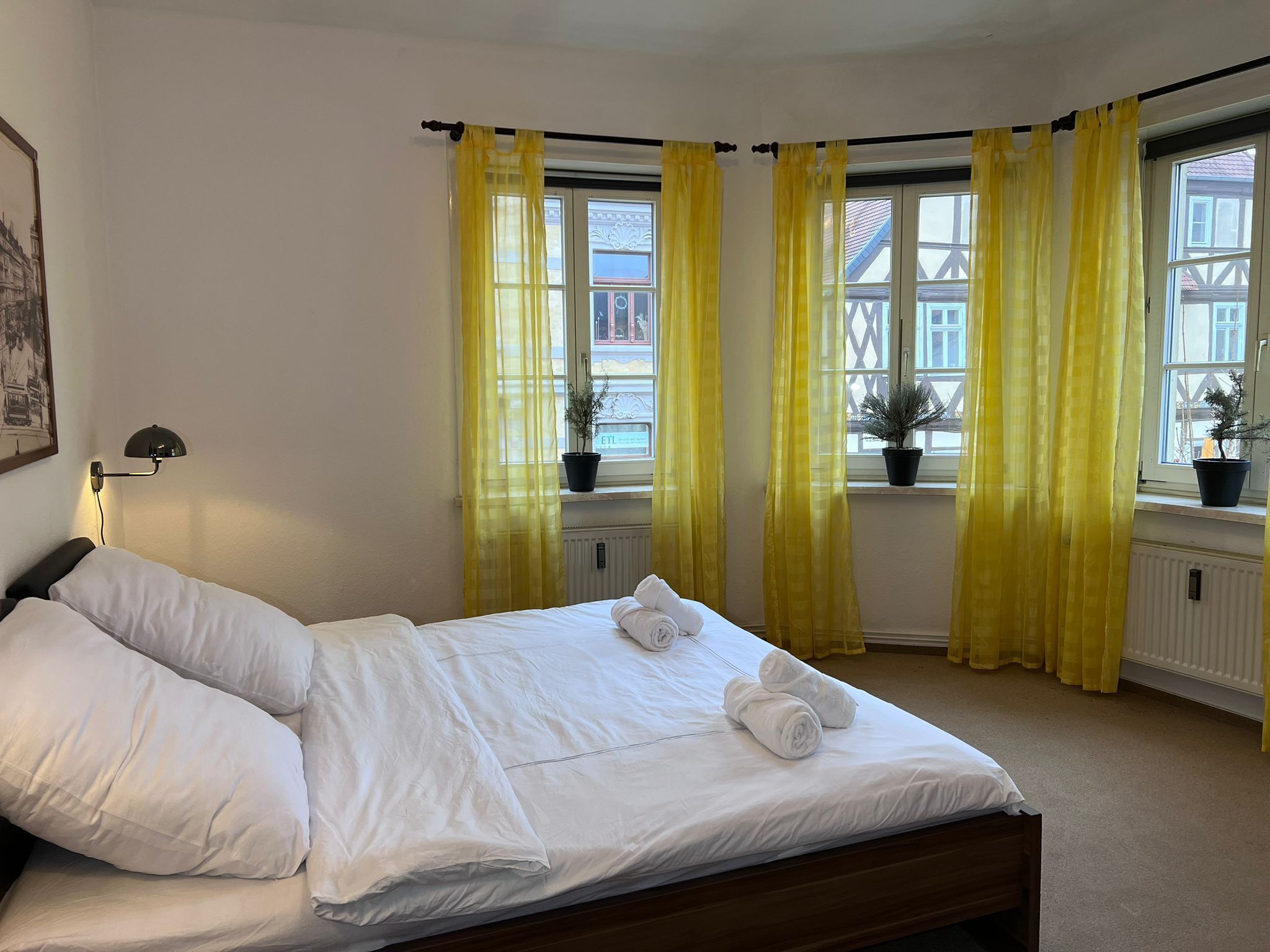 Ein ordentlich bezogenes Bett mit weißen Leinen und Handtüchern, eine schwarze Schreibtischlampe und ein Fenster mit gelben Vorhängen.