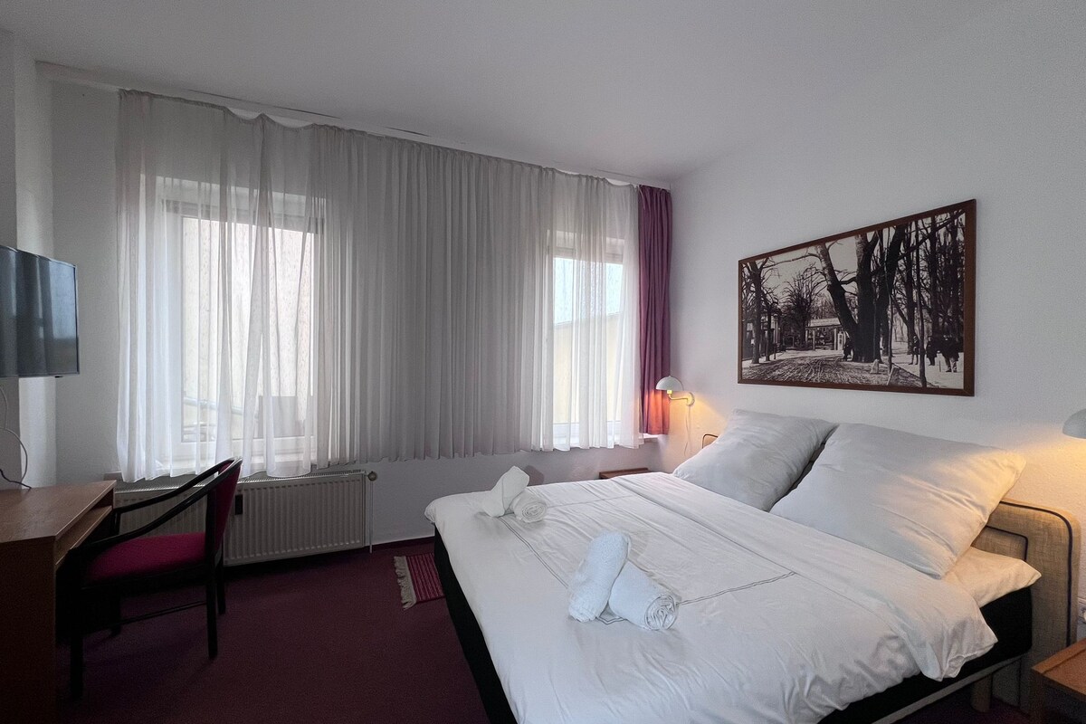A hotel room with a bed, a desk, a chair, a TV, and a painting on the wall.
