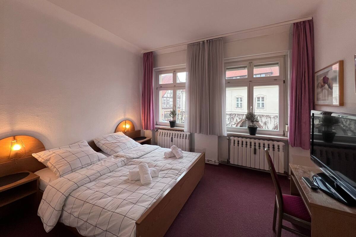 A hotel room with a bed, bedside lamps, a TV, a window with curtains, and a radiator.