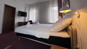 A hotel room with a bed, a desk, a chair, a TV mounted on the wall, and a window with curtains. - Hotel Doko Gardelegen (Gardelegen)