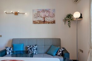 A modern living room with a grey sofa, two patterned cushions, a glass coffee table, a wall-mounted light fixture, a painting of a tree, a floor lamp, and a potted plant. - Adele's House - with pool terrace and WiFi (Sirmione)