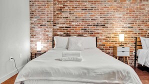 Una cama en una habitación con pared de ladrillo, una mesita de noche y un velador.