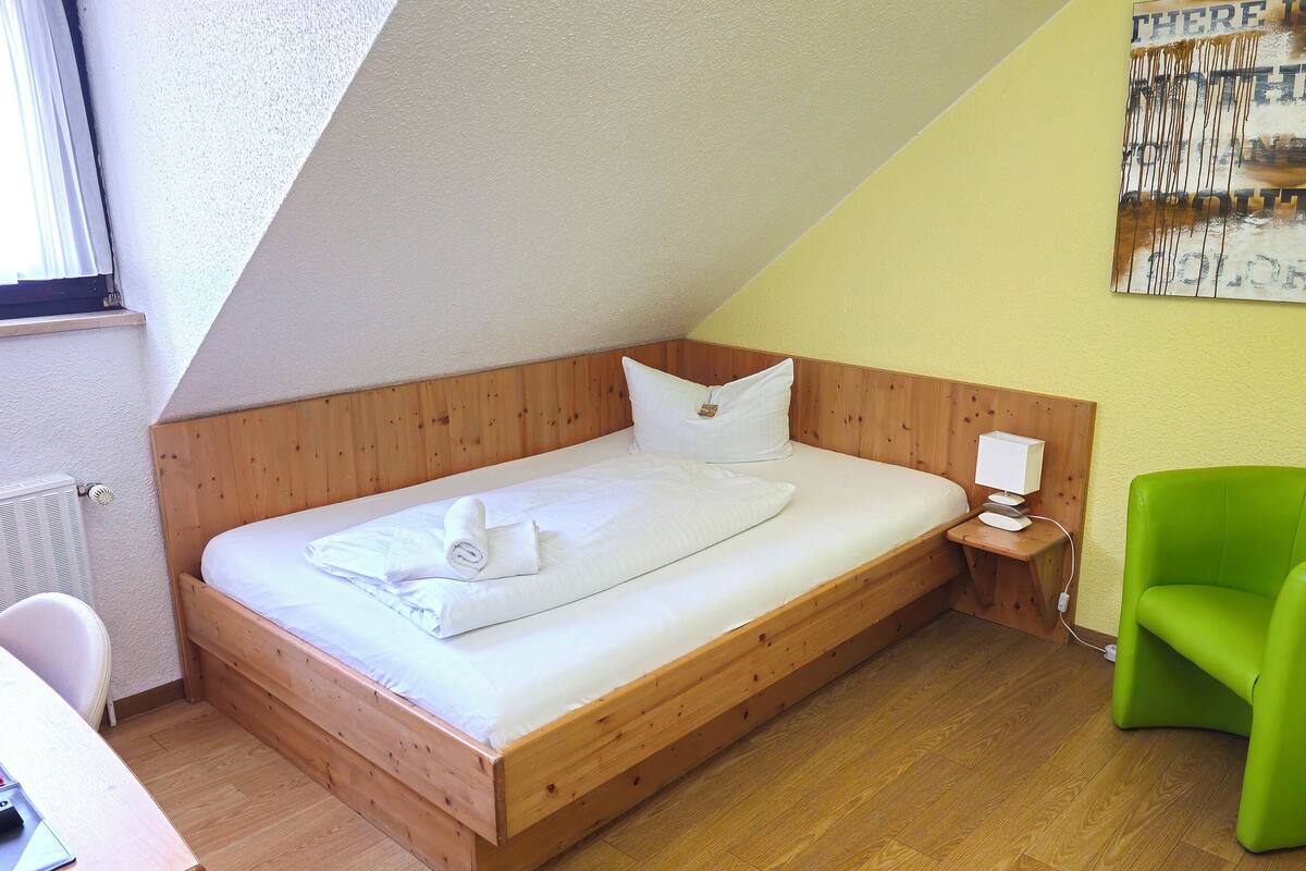 Un lit simple avec une tête de lit en bois, une table de chevet avec une lampe, une chaise verte et un tableau accroché au mur.