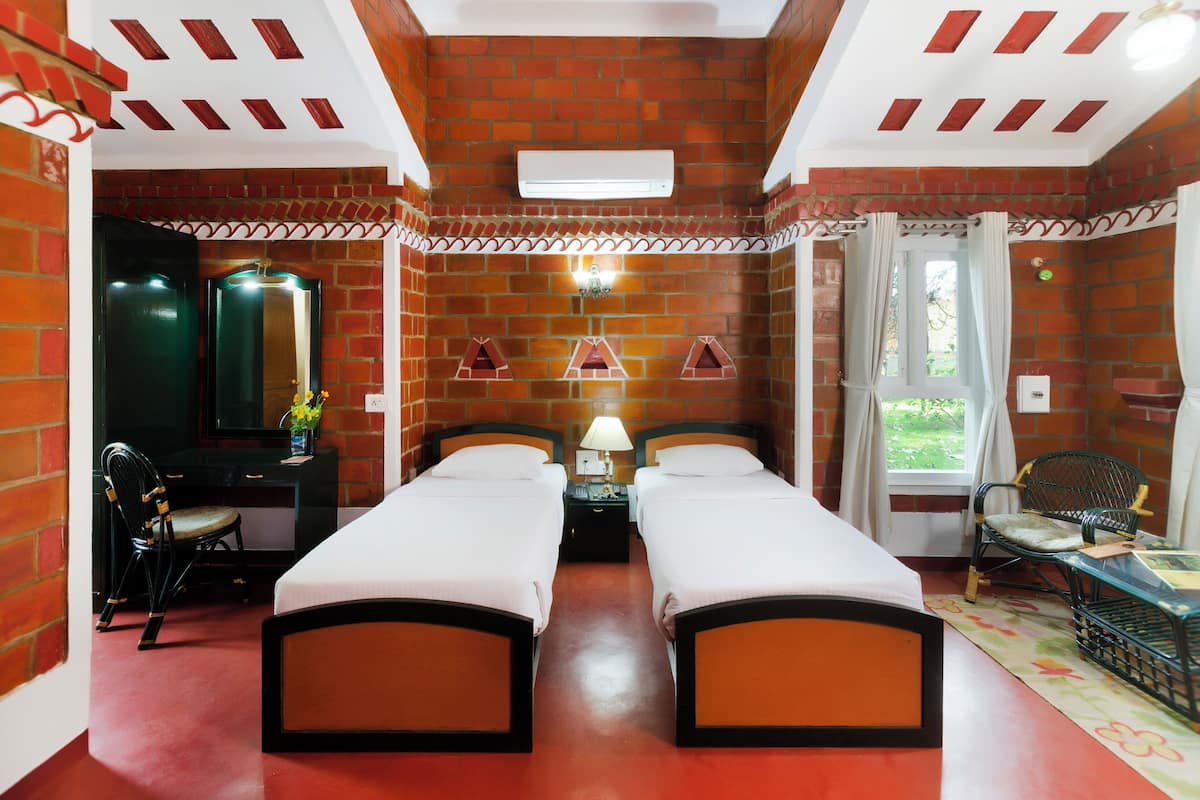 A room with two beds, a desk with a chair, a window with curtains, and a brick wall.