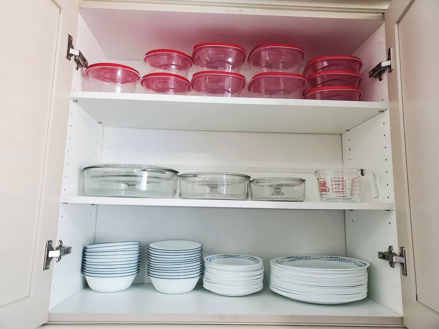A kitchen cabinet with three shelves. The top shelf has red containers, the middle shelf has clear glass containers, and the bottom shelf has stacked white plates.