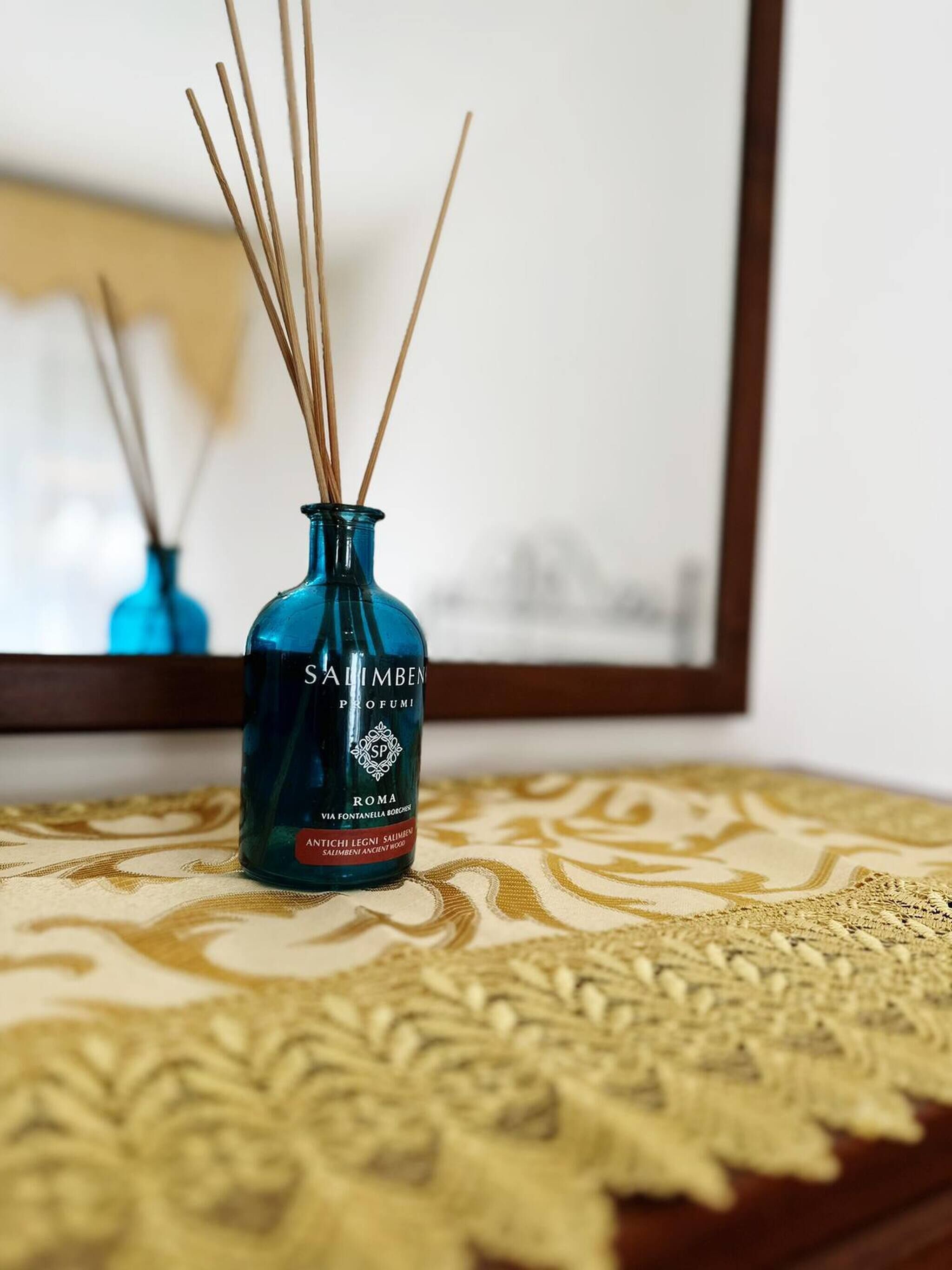 A blue glass bottle with reed sticks on a patterned tablecloth, placed in front of a mirror.