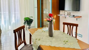 A dining area with a table set for four, a vase with flowers, a television mounted on the wall, and a window with sheer curtains. - Ti Aspettiamo nel Nostro Luminoso Appartamento (Gela)
