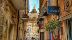 A narrow street with buildings on both sides, a sign for 'barocco', and a distant church tower. - Ti Aspettiamo nel Nostro Luminoso Appartamento (Gela)