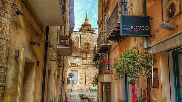 A narrow street with buildings on both sides, a sign for 'barocco', and a distant church tower. - Ti Aspettiamo nel Nostro Luminoso Appartamento (Gela)
