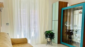 A living room with a beige sofa, a potted plant, a glass cabinet, and a window with sheer curtains. - Ti Aspettiamo nel Nostro Luminoso Appartamento (Gela)