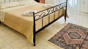 A neatly made bed with a beige bedspread and pillows, a black metal headboard, a bedside table with a lamp, and a patterned rug on a tiled floor. - Ti Aspettiamo nel Nostro Luminoso Appartamento (Gela)