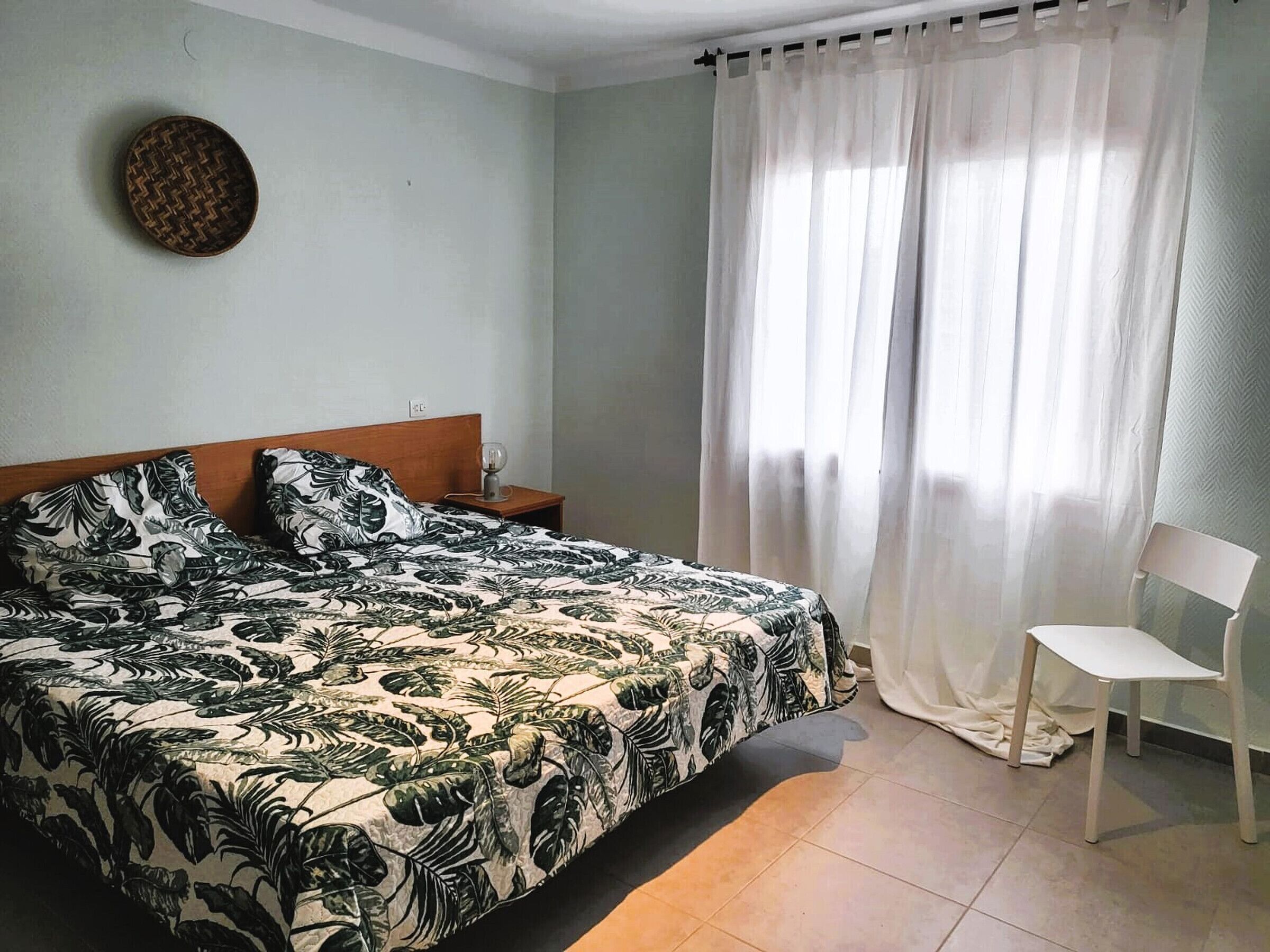 Un dormitorio con cama, mesita de noche, silla y ventana con cortinas.