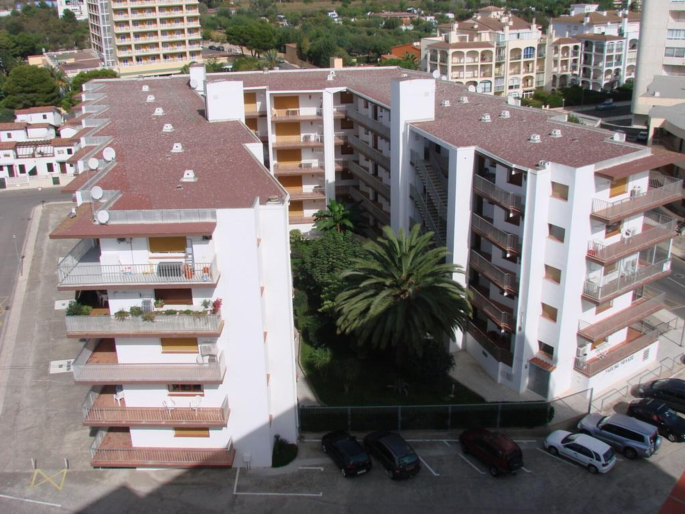 A multi-story residential building with balconies, a parking area, and a palm tree.