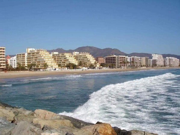 A coastal hotel with multiple floors, a beach in the background, and mountains in the distance.