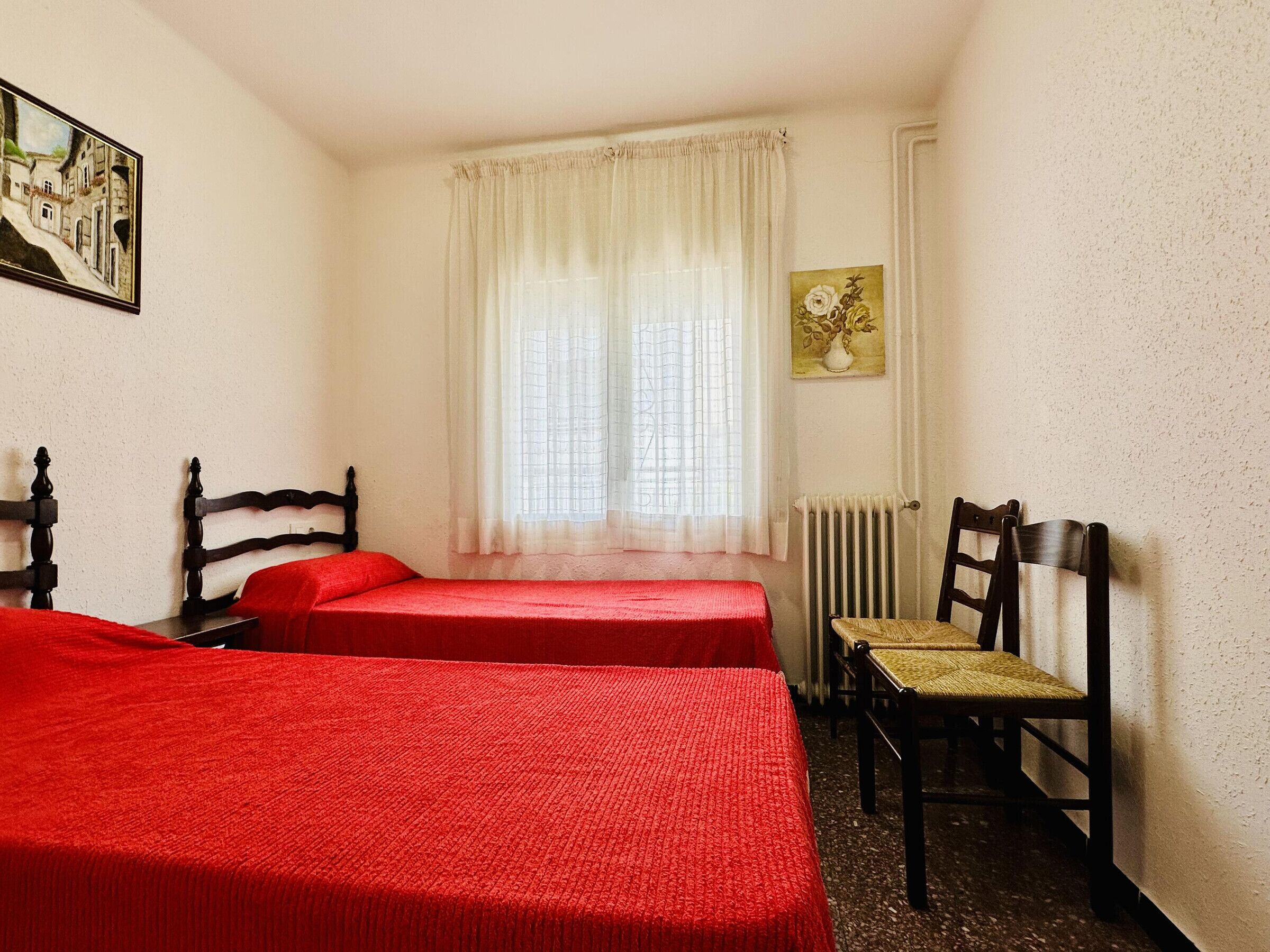 Une petite pièce simple, avec deux lits simples, une chaise, un radiateur et une fenêtre avec des rideaux.