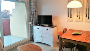 Una habitación pequeña y bien iluminada, con un ventanal corredero que da a un espacio exterior, un mueble blanco con un televisor de pantalla plana, una mesa de madera con sillas y una lámpara de pared.