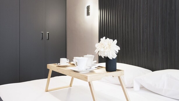 A neatly made bed with white linens, a bedside table with a white vase and tea set, and a dark wooden panel wall. - Easylife -Modern apartment with Patio C.Sforzesco (Milano)