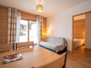 Um quarto acolhedor com uma mesa de jantar de madeira, um sofá e uma cama. Há uma janela com cortinas, uma luminária na parede e uma porta que dá para outro quarto.