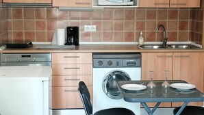 A compact kitchen with a microwave, coffee maker, dishwasher, and a small dining area with a table and chairs. - Charming Apt in Heart of Santa Margarida, Near Beach, Terrace, & Parking (Roses)