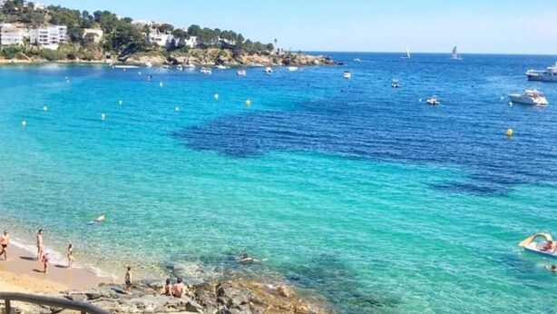 Cena costeira com águas azul-claras, barcos e uma praia com pessoas.