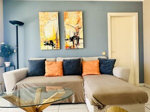 Ein Wohnzimmer mit einer Couch, einem Glastisch, zwei abstrakten Gemälden, einer Stehlampe und einer Topfpflanze.