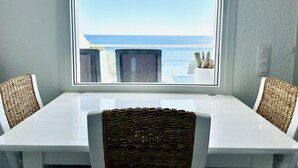 Ein Essbereich mit Blick auf den Ozean durch ein Fenster, mit einem weißen Tisch und Korbstühlen.