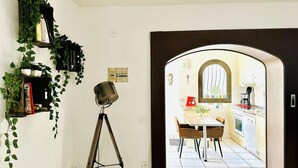 A room with a tripod-mounted lamp, a dining area with a table and chairs, and an archway leading to a kitchen with a window and appliances. - Charming House Near Natural Park - Hiking, Biking, Windsurfing & Communal Pool (Roses)