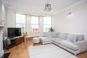 Un salon comprenant un canapé d’angle, un téléviseur à écran plat, un meuble de rangement en bois et un tapis à motifs.