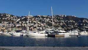 Uma marina com vários iates atracados, um píer de madeira e um morro com edifícios residenciais ao fundo.