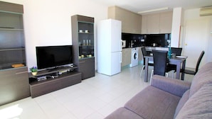 Uma sala de estar moderna com televisão de tela plana, sofá, área de refeições com mesa e cadeiras, cozinha com máquina de lavar e um pequeno armário com portas de vidro.