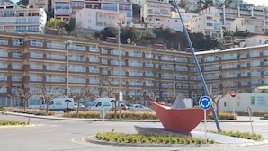 Um edifício residencial com vários andares e varandas, uma rotatória com uma escultura vermelha e uma placa de sinalização indicando zona proibida.