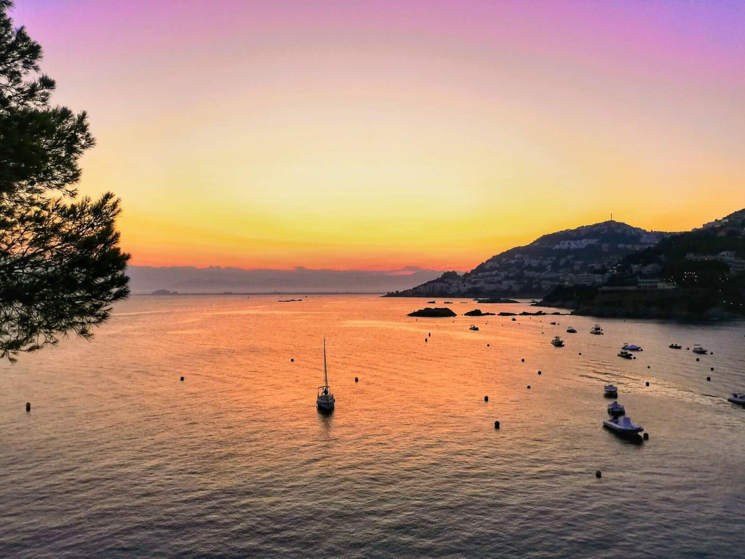 Une vue sur la côte, avec des bateaux, un voilier et un paysage montagneux au coucher du soleil.