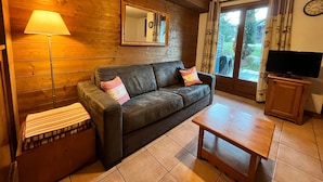 A cozy living room with a wooden wall, a sofa with cushions, a lamp, a television on a wooden stand, and a sliding glass door leading to an outdoor area.