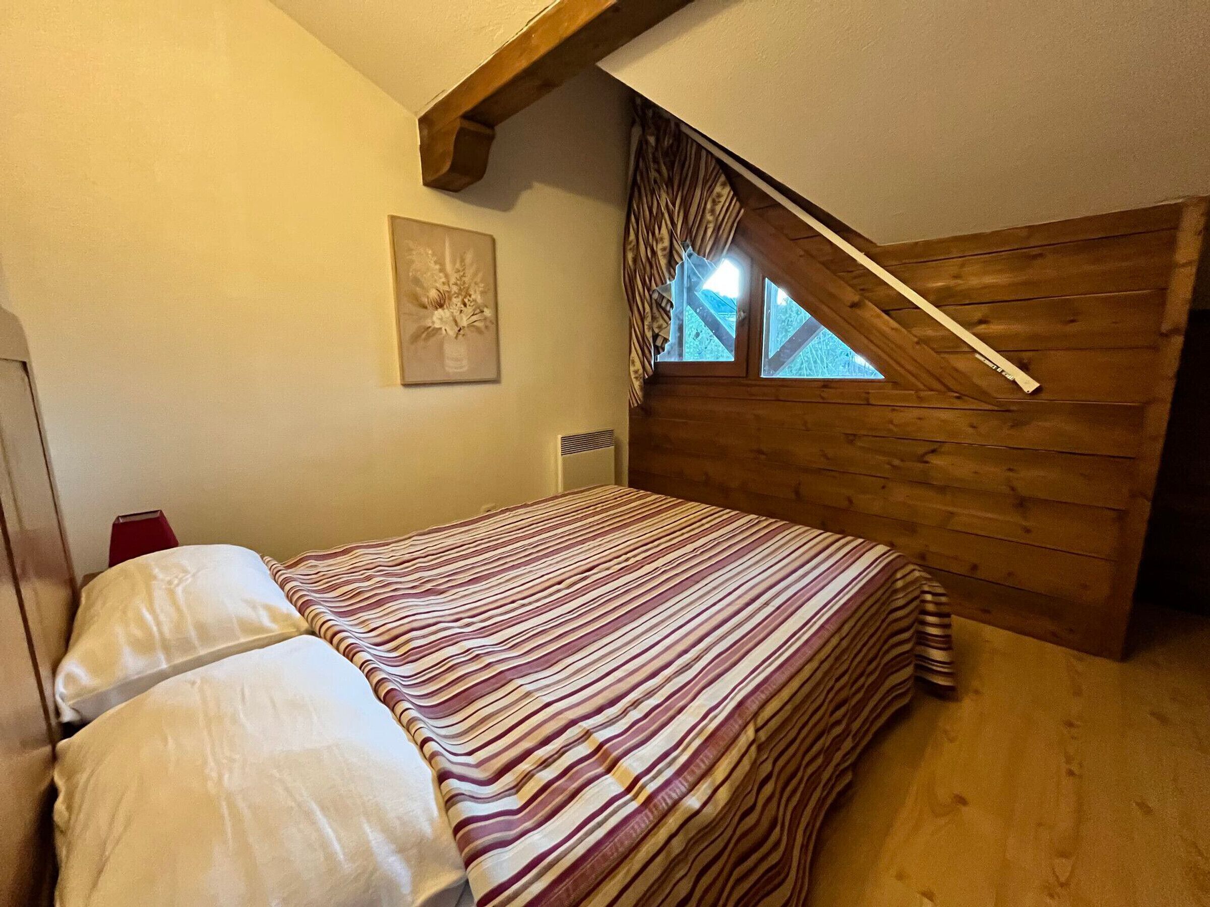 A bedroom with a sloped ceiling, a bed with striped bedding, a wooden headboard, a window with curtains, and a wall-mounted picture.