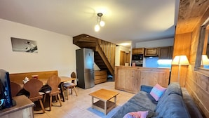 A cozy living room with a sofa, coffee table, and wooden dining table. A kitchen area with a microwave and cabinets is visible in the background.