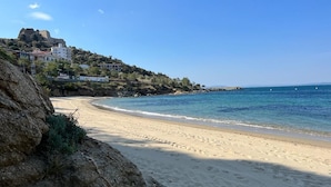 Uma praia arenosa com um afloramento rochoso à esquerda e um edifício numa colina ao fundo.