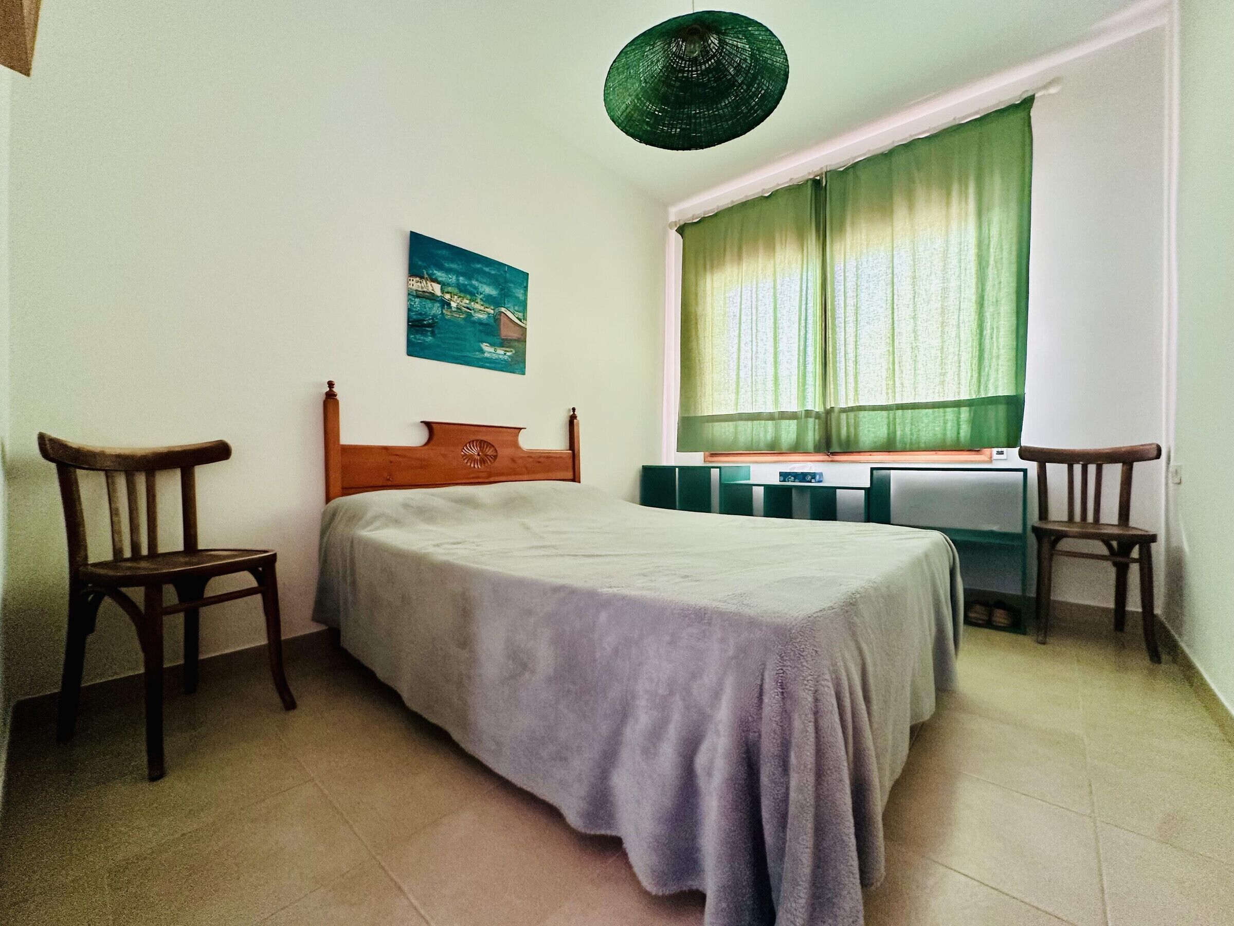 A bedroom with a bed, two chairs, a desk, a window with curtains, a wall-mounted picture, and a hanging light fixture.